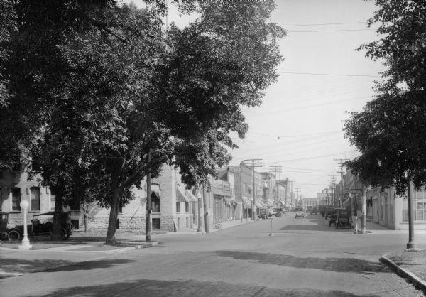 View down street toward storefronts on either side of a dirt road in the business district. Automobiles are parked on both sides of the street, and two automobiles and a horse-drawn carriage are moving along the street. A sign on the right advertises a "Garage."