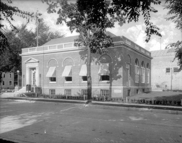 View across street toward the facade and right side of the post office. The main entrance, set to the left, is framed with a neoclassical surround. There are decorative arches above the windows, and striped awnings protect the interior from direct sunlight.