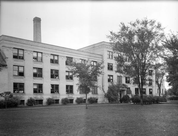 Exterior view across lawn toward Northwestern College. Young men are posing in many of the windows. The building features a neoclassical style, with a columned porch and  decorative brickwork.