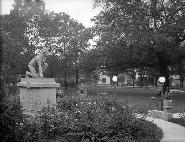 A sculpture of a man kneeling stands in the middle of a park surrounded by plants and a sidewalk. In the background is a lawn and trees, and a street lined with houses.