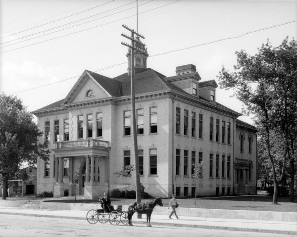 View across street toward the facade and right side of a high school building. A postman walks along the sidewalk in front, and nearby a man sits in his horse-drawn carriage.