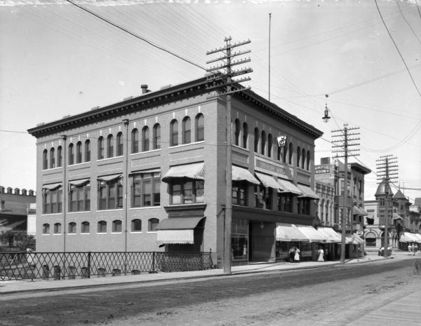 Angled view across street toward a railing along a sidewalk, and the front facade and left side of a Masonic temple building. The ground floor contains a store. Awnings cover the windows on the ground and second floor. People, mostly women are walk down the sidewalk.