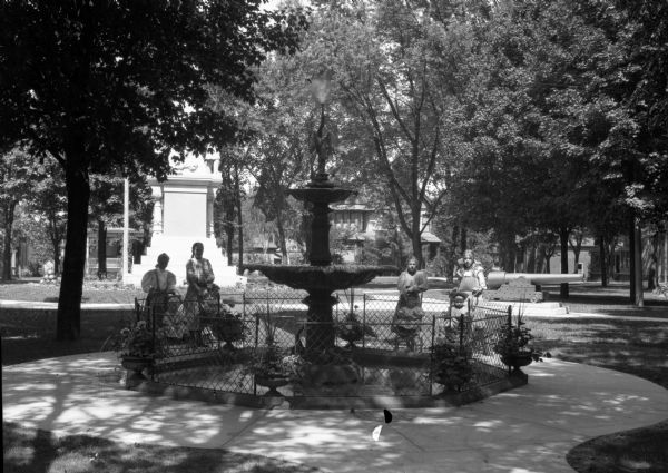 Four girls are each standing behind a child sitting in a stroller while posing around Lewis Fountain in Veteran's Park. Houses and the Civil War Veteran's Memorial are in the background.