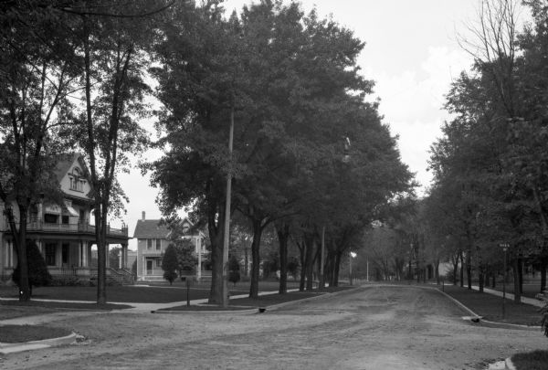 View toward intersection of a tree-lined unpaved street. Two large houses wide wide lawns are on the left.