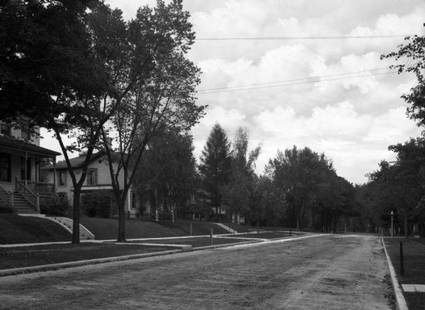 View across street towards houses on the left. A wide terrace planted with trees is along the sidewalk. Steps lead up a small rise on the front lawns of the houses.