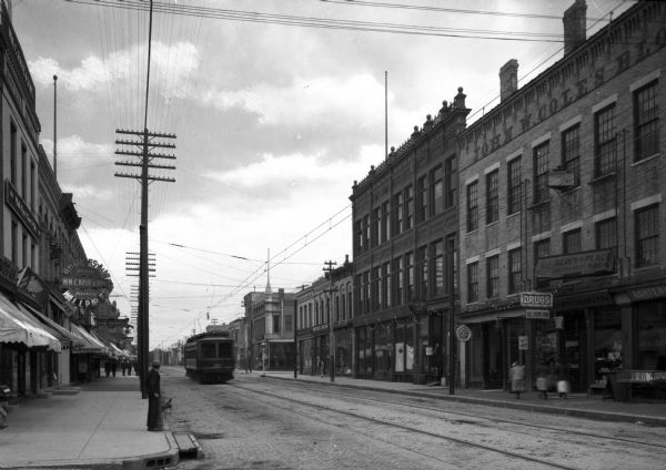 View along left side sidewalk toward shops and businesses lining a commercial street. A large sign outside a shop on the left reads: "Wholesale Wallpaper, Wm. C. Raue & Sons." A storefront on the right has signs for "Drugs" and "Ice Cream Soda." A streetcar is moving up the street on the left.