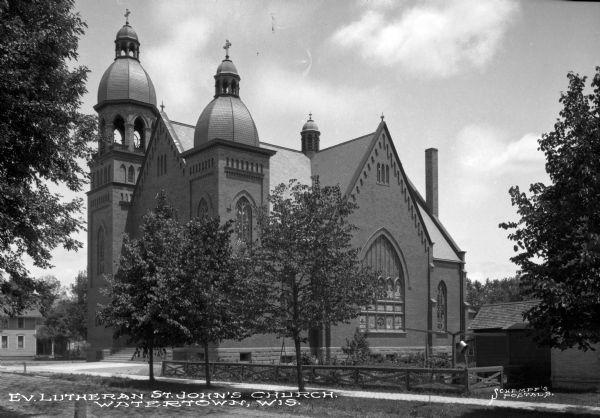 View across lawn and sidewalk toward the facade and left side of St. John's Evangelical Lutheran Church, partially obscured by trees. The facade has two domes on each side of the main entrance, the left being a bell tower. There are large stained glass windows. Caption reads: "Ev Lutheran St. John's Church, Watertown, Wis."