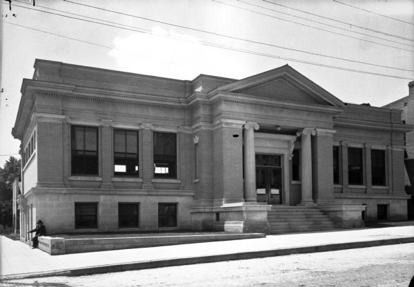 View across street toward the facade of the Watertown Public Library. The Neoclassical style informs elements of the building, including the porch, pediment, and the columns. An older man with a cane sits on a retaining wall at the left side of the structure.
