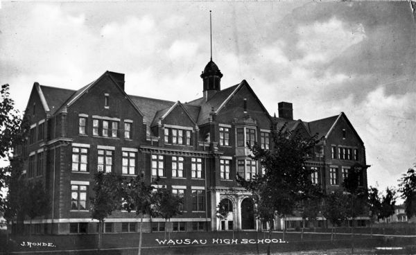 View toward the Wausau High School facade, with an stone arched entrance and central stone bay windows above. The flag pole sits on top of the tower. Caption reads: "Wausau High School."