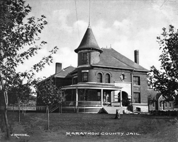View across lawn toward the facade and side of the Marathon County Jail. A porch wraps around one corner of the first floor, below a round turret that extends to the third floor. A stone archway marks the main entrance. Caption reads: "Marathon County Jail."