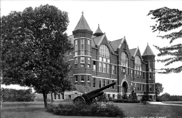 A view of Main Hall at St. Norbert College.  Turrets and stone decor accent the brick building and a cannon surrounded by shrubbery sits on the lawn.