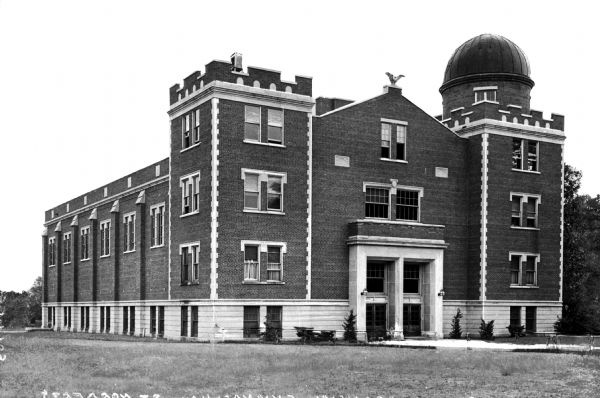 A view of the facade and side of the brick gymnasium at St. Norbert College. Stone is used for the basement floor and in decorative elements. This building also serves as the observatory, seen on the tower to the right. An eagle statue tops the building above the entrance.