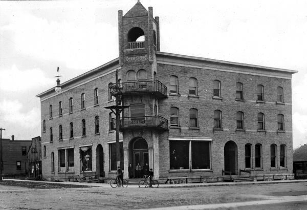 View of Hotel Hutchinson, situated at the corner of two streets. The entrance stands beneath a bell tower and a group of boys, some with bicycles, stands near the doorway. The stone marker beneath the bell reads, "1899."