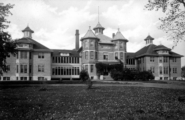 View across lawn the Waupaca County Asylum facade. The structure consists of three buildings connected by windowed passageways. The building is symmetrical, each topped with a ventilation cupola on top of the roofs. The middle building has two polygonal turrets, which are mirrored in the bay windows on the flanking buildings.