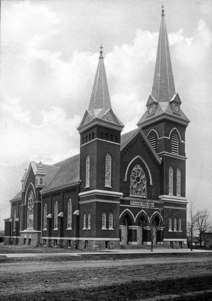 View across road toward the front facade and left side of St. Peter's Evangelical Lutheran Church. The Gothic Revival-style structure has two steeples, the larger to the right housing the belfry. Three doors form the main entrance and every visible window is made of stained glass.