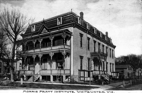 Veew toward the facade and right side of the Morris Pratt Institute on a street corner. The roof line has an ornate iron railing running around its perimeter and the railings on the porch and balcony are elaborately decorated. Caption reads: "Morris Pratt Institure, Whitewater, Wis."
