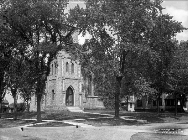 A view of the facade of an Episcopal church, partially obscured by trees. A house stands to the building's right.
