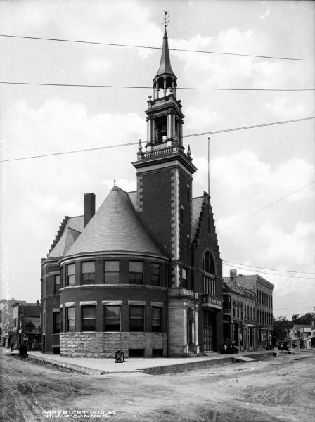 A view of the facade and side of City Hall. The main entrance is part of the bell tower. Men are sitting on the bench outside to the right.