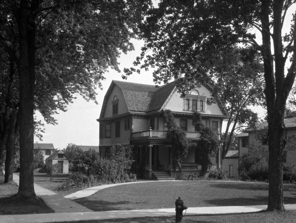 View of a house and yard as seen from the street.  The gables of the house suggest Dutch Colonial style.  Large plants line the walkway and vines trail up the porch columns. A small shed or outhouse, decorated with the same shingles as the house, is on the left.