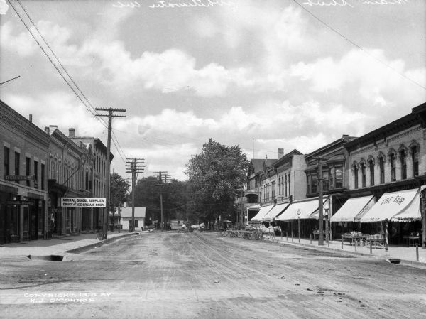 A view of businesses lining Main Street. Signs on the buildings to the left advertise "Dentist," "School Books," and "Normal School Supplies. Drugs and Ice Cream Soda." The awning to the right reads, "The Fair."