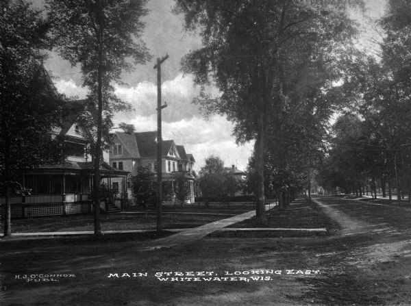 A view looking east along the left side of a tree-lined Main Street with houses on the left. Caption reads: "Main Street, Looking East, Whitewater, Wis."