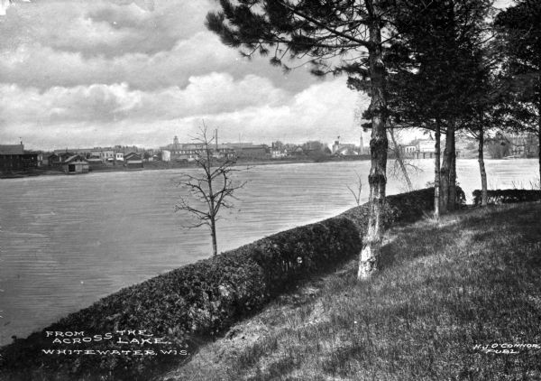 View from shoreline across the river toward the town of Whitewater on the opposite riverbank. Caption reads: "From Across the Lake. Whitewater, Wis."