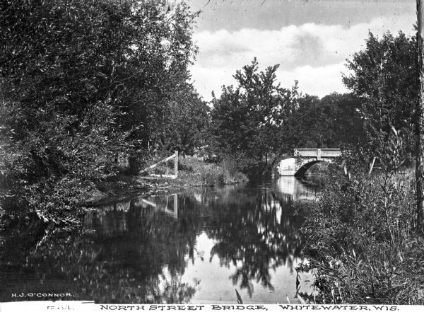 View upriver toward the bridge. A wooden structure is to the left of the bridge. Caption reads: "North Street Bridge, Whitewater, Wis."