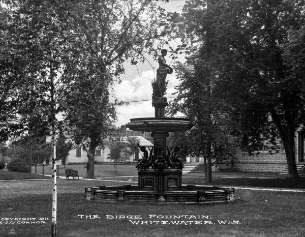 View of the Neoclassical-style Birge Fountain. Mythical figures decorate the foot of the structure and a woman pouring water from a basket tops the fountain. Below her are unclothed children sitting beside fish. Caption reads: "The Birge Fountain, Whitewater, Wis."