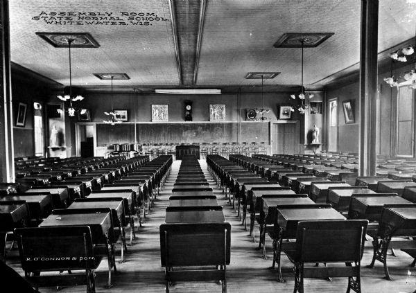 A view of the interior of the assembly room at the State Normal School, looking toward the front of the room.  Long rows of desks fill the room.  Above the chalkboard are bas relief panels, a clock, a photograph and painting.  Classical sculptures stand in the corners of the room. Caption reads: "Assembly Room, State Normal School, Whitewater, Wis."