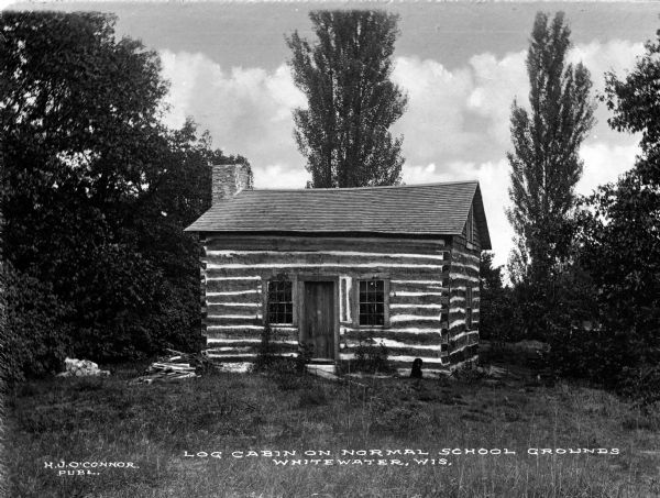 A view of the facade of a log cabin on the grounds of the State Normal School. A black dog sits in front of the building. Caption reads: "Log Cabin on Normal School Grounds, Whitewater, Wis."