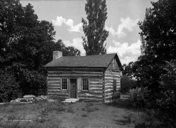 A view of a cabin on the grounds of the State Normal School.  A pile of logs sits to the left.