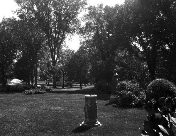 A view of a lawn at the State Normal School accented with a bust and sundial. Trees and various plantings dot the landscape.