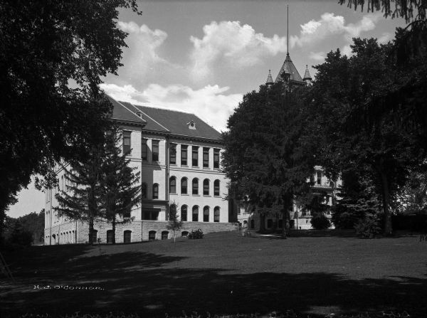 A view of the facade of the State Normal School. The middle section, including a bell or clock tower, is mostly obscured by trees.