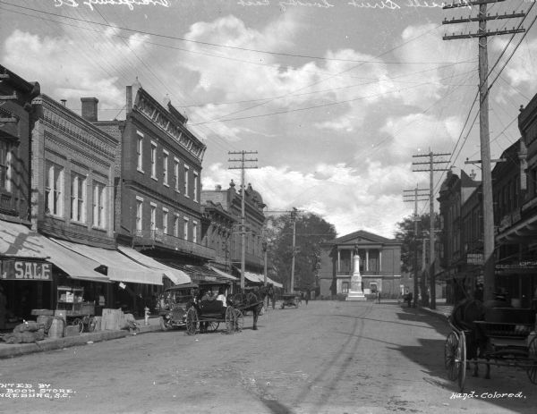 View of horse-drawn vehicles and automobiles on a street. A banner partially reading "SALE" is on a storefront.
