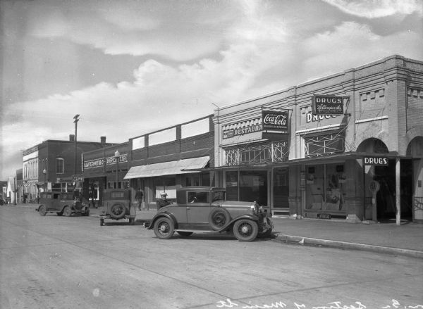 View of several cars parked along Main Street. Business signs read: "Hollingsworth's Drugs," "Harter Motor Company," "American Cafe" and "Coca-Cola."