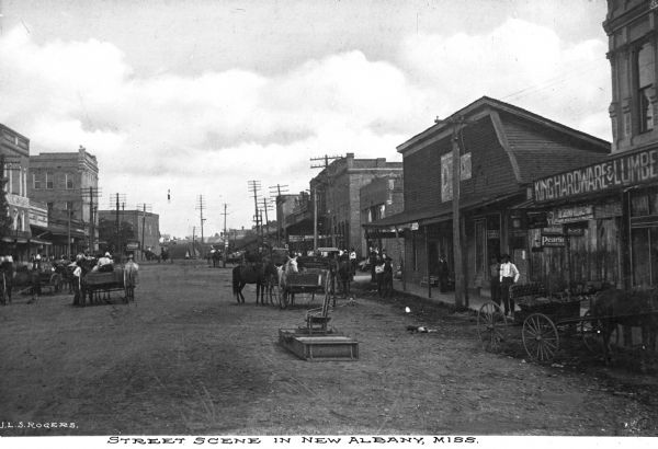Signs read: "King Hardware and Lumber" "Mayors office" "W.N. Parks" "Fish Bros. Wagon Co." and "Coca-Cola." Caption reads: "Street Scene In New Albany, Miss."