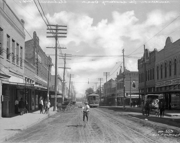 View of a boy riding a bicycle in a city street with an electric cable car on tracks in the background. Horse-drawn carts are visible as well. Business signs read: "American dry goods and shoes, millinery" "The Model Clothing Co." "Foster-Fain Drug Co." "Cyrus and Cyrus" and "Dr. B.H. Turner office."