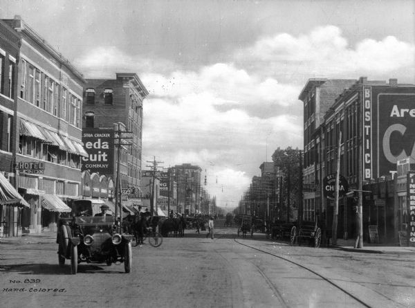 View of a busy street, lined with businesses, pedestrians, automobiles and carts. Many business signs are obscured, but some read: "Hotel Lenox," "Thomason's Cafe," "Hotel Waverly," "J.C. Redfield Prop. Pure Food Grocery," "Bosticb" and "Joe's Quick Lunch."