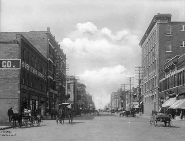 View down a street lined with businesses and full of horse-drawn carts. Business signs read: "Hooker-Hendrix Hardware Co.," "Light & Power," "Norman Hotel," "Dr. McCollister Sanitarium Specialist" and "Oklahoma Brokerage Co."