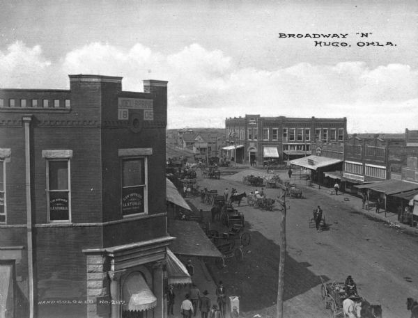 Elevated view of a wide city street. Horse-drawn carts are in the street and pedestrians crowd the sidewalks. Sign in window reads: "Law Offices of D.A. Stovall." Sign on top of building reads: "Joel Spring 1805." Caption reads: "Broadway 'N' Hugo, Okla."