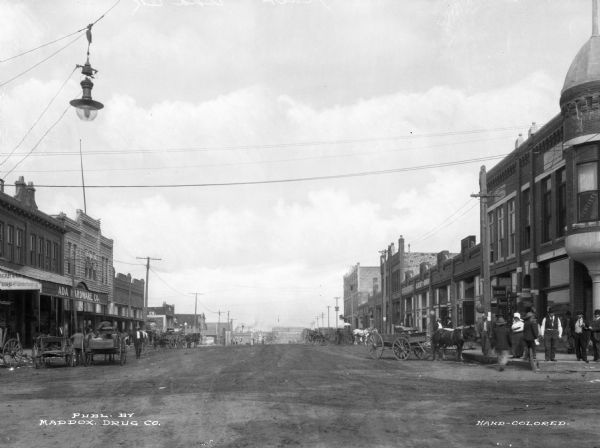 Main street lined with storefronts, horse-drawn carts and pedestrians. Electric lights hang over the street. Business signs read: "Madox Drug" "Guest Bros." and "Ada Hardware Co. Furniture and Undertaking." Text on photograph reads: "Publ. by Maddox Drug Co."