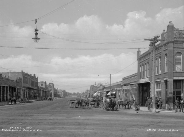 Main street lined with storefronts, horse-drawn carts and pedestrians. Electric lights hang over the street. Signs read: "Ada National Bank" "Dr. Thompson Dentist" "Harris Drugs." Text on photograph reads: "Publ. by Maddox Drug Co."