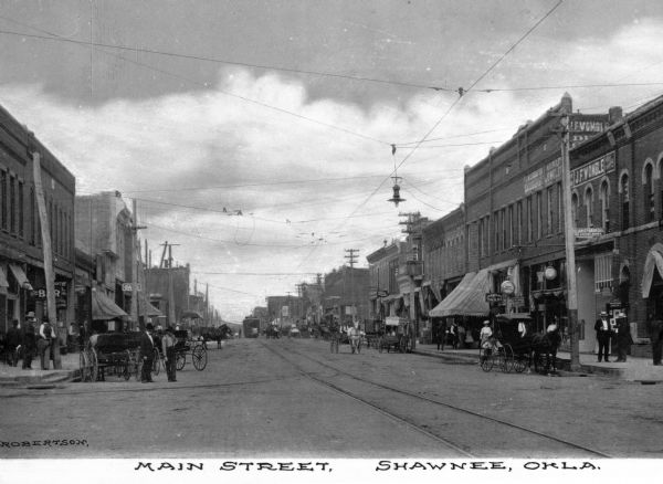 Main street lined with storefronts, horse-drawn carts and pedestrians. Electric lights hang over the street. Business signs read: "J.H. Kreiling Tailor" "J.F. Womble" "Schloss." Caption reads: "Main Street, Shawnee, Okla."