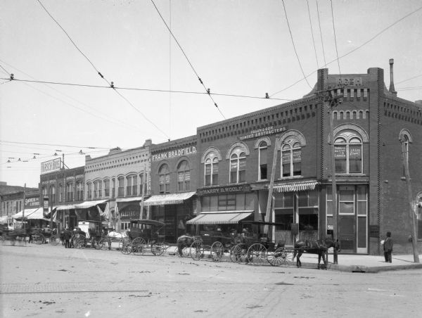 North side view of the town square with storefronts, horse-drawn carts and pedestrians. Electric power lines hang over the street. Business signs read: "Fagan and Son. Bonded Abstracters" "J.A. Lee Attorney" "Hudkins Real Estate & Loan Co." "W.A. Graves Attorney" "King and Pratt Real Estate and Loans" "Harry B. Woolf The Man Store" "Frank Bradfield Real Estate Loans" "Drugs and Jewelry" "Electric Theatre" "Gannon & Goulding" "Hirsch Bros." "The Fitwell Shoe Co." and "W.C. Plaeffle Diamonds."