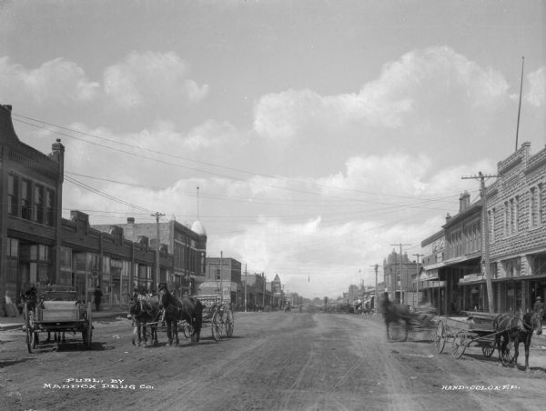 Main street lined with storefronts, horse-drawn carts and pedestrians. Electric power lines hang over the street. Business signs read: "Case & Hudelson Furniture and Stoves" "Ada Hardware" "Peter's Shoes" "Guest Bros. Dry Goods." Text on photograph reads: "Publ. by Maddox Drug Co."