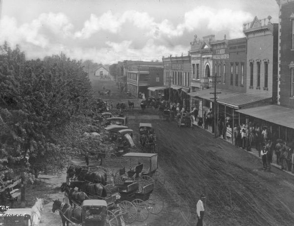 Elevated view of First Street lined with trees, storefronts, horse-drawn carts and pedestrians. Text on buildings reads: F.Z. Meeks Wholesale Grocery Co." and "Benton Co. Hdw. Co."
