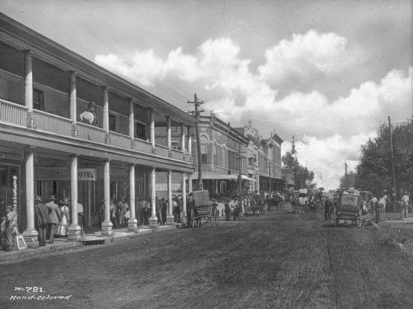 First street lined with trees, storefronts, horse-drawn carts and pedestrians.