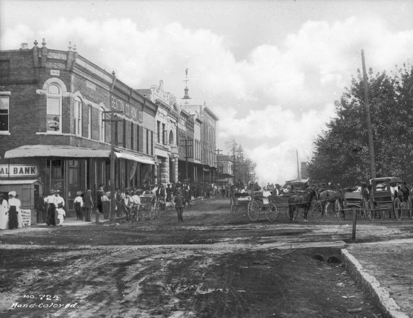 First street lined with trees, storefronts, horse-drawn carts and pedestrians. Text on building reads: "Benton Co. Hdw. Co." Sign in window reads: "Dr. Curry's Office."