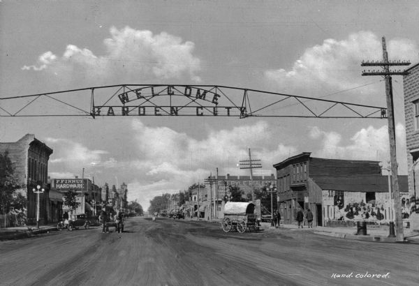 View down Main Street with overhead sign reading "Welcome Garden City" sign. Text on buildings reads: "E.J. Pyle Real Estate" and "F. Finnup. Hardware." Circus advertisements are on the right side of the street.