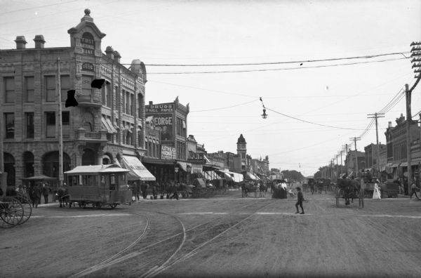 Street lined with storefronts, horse-drawn carts, an electric cable car and pedestrians. Electric power lines hang over the street. Business signs read: "First National Bank," "Brown Sisters Millinery" and "Austin Black Hardware Co."